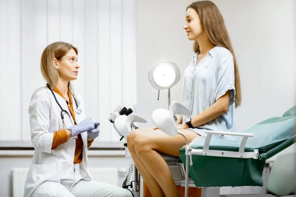 A woman seated in an examination room engages in conversation with a doctor in a clinical setting.