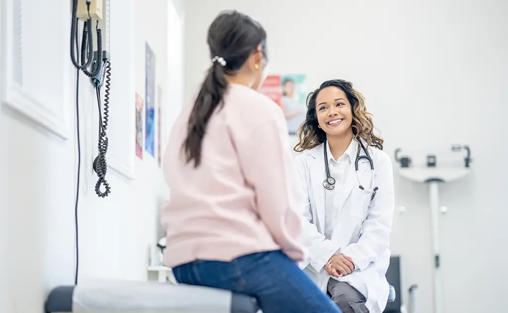 A female doctor converses with a patient in a clinical setting, discussing health concerns and treatment options.